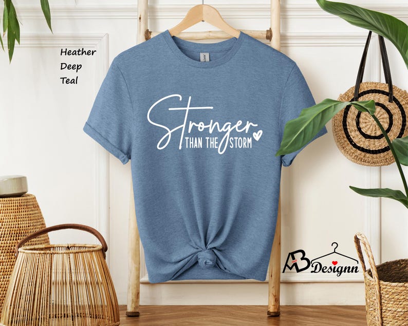 Może przedstawiać: Koszulka w kolorze wrzosowo-ciemnozielonym z napisem "Stronger than the storm" w białym kolorze z małym sercem.