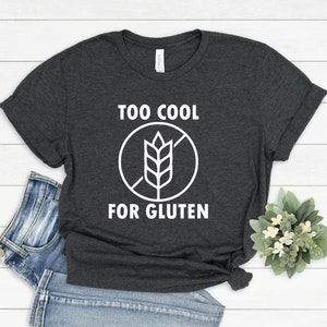 May include: A dark gray t-shirt with white text that reads "Too Cool For Gluten" and a white graphic of a wheat stalk with a diagonal line through it.