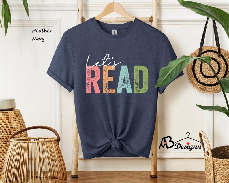 May include: A heather navy blue t-shirt with the text "Let's Read" in a colorful, distressed font. The text is in shades of pink, blue, green, and yellow.