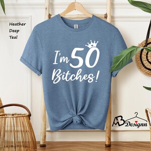 May include: A heather deep teal short-sleeve t-shirt featuring white screen-printed text that reads I'm 50 Bitches! with a small crown graphic above the number 50. The shirt has a crew neckline and is displayed on a wooden hanger.