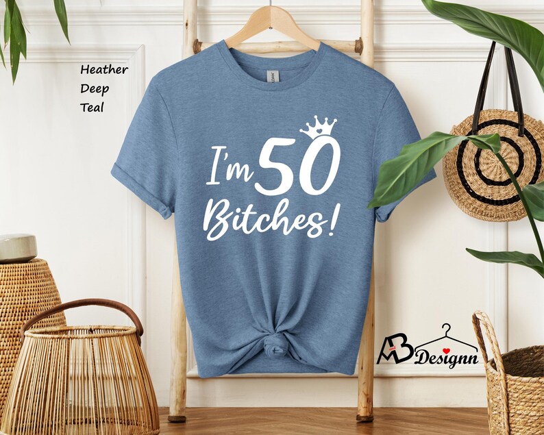 May include: A heather deep teal short-sleeve t-shirt featuring white screen-printed text that reads I'm 50 Bitches! with a small crown graphic above the number 50. The shirt has a crew neckline and is displayed on a wooden hanger.