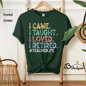 Może przedstawiać: Leśna zielona koszulka z napisem "I CAME. I TAUGHT. I LOVED. I RETIRED. #TEACHERLIFE" w kolorowych literach. Koszulka wisi na drewnianym wieszaku. Koszulka ma okrągły dekolt i kr&oacute;tkie rękawy.