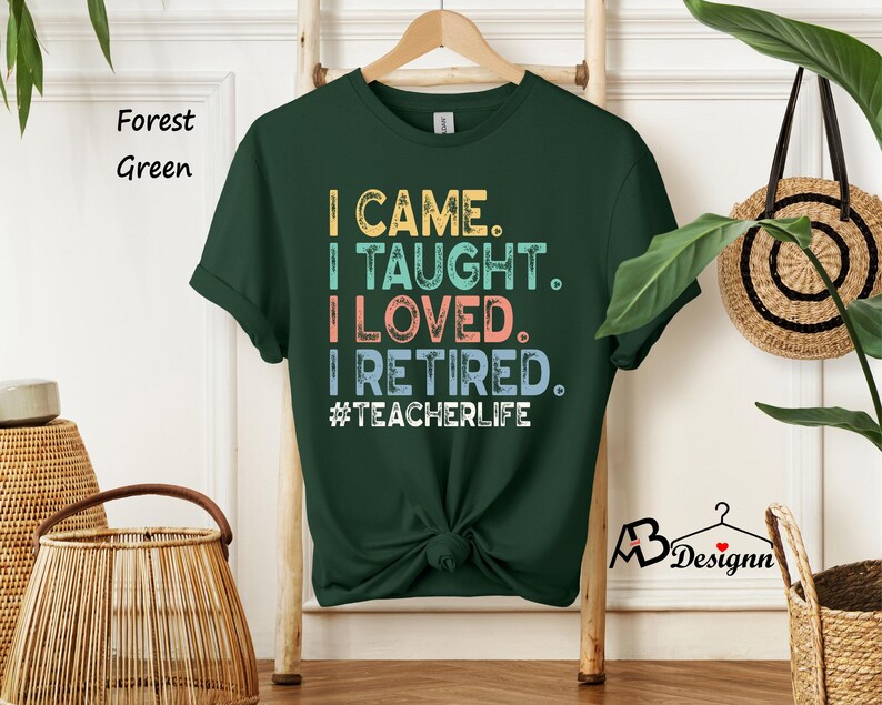 Może przedstawiać: Leśna zielona koszulka z napisem "I CAME. I TAUGHT. I LOVED. I RETIRED. #TEACHERLIFE" w kolorowych literach. Koszulka wisi na drewnianym wieszaku. Koszulka ma okrągły dekolt i kr&oacute;tkie rękawy.