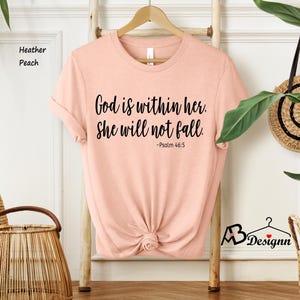 Peut inclure: Un t-shirt couleur p&ecirc;che avec le texte "Dieu est en elle. Elle ne tombera pas. -Psaume 46:5" imprim&eacute; en noir.