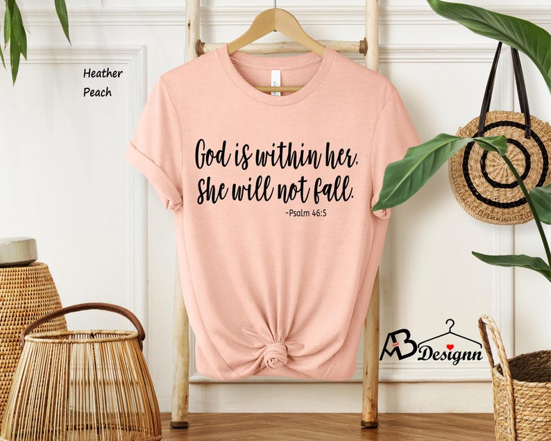 Peut inclure: Un t-shirt couleur p&ecirc;che avec le texte "Dieu est en elle. Elle ne tombera pas. -Psaume 46:5" imprim&eacute; en noir.