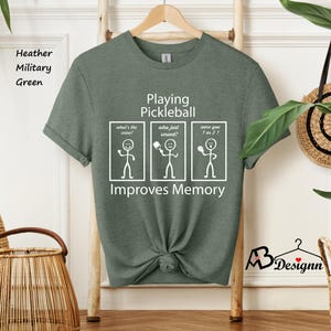 May include: A green t-shirt with the text "Playing Pickleball Improves Memory" and a stick figure illustration. The illustration shows three stick figures with thought bubbles that say "What's the score?", "Who just served?", and "Were you 1 or 2?"