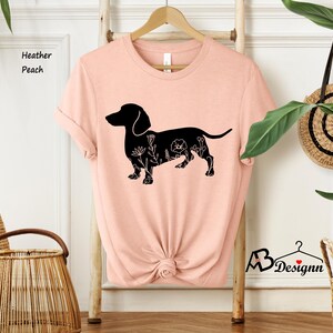 May include: A peach coloured t-shirt featuring a black silhouette of a dachshund with floral details. The text "Heather Peach" is visible in the top left corner of the image.