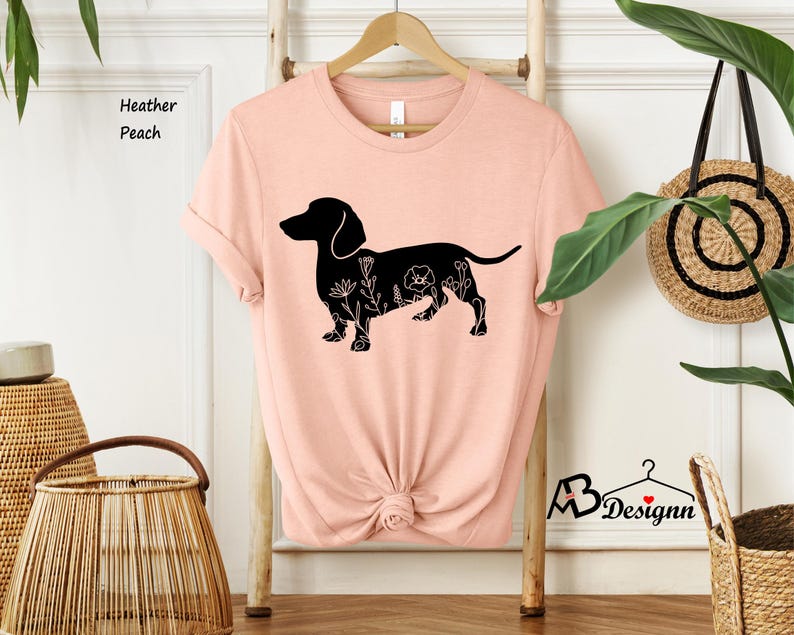 May include: A peach coloured t-shirt featuring a black silhouette of a dachshund with floral details. The text "Heather Peach" is visible in the top left corner of the image.