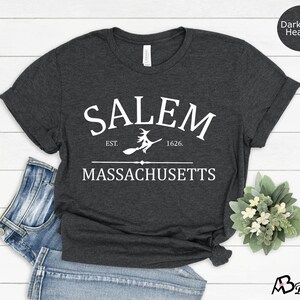 May include: Dark grey heather t-shirt with white text that reads "SALEM EST. 1626. MASSACHUSETTS" with a witch flying on a broomstick graphic.