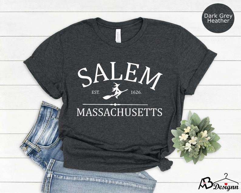 May include: Dark grey heather t-shirt with white text that reads "SALEM EST. 1626. MASSACHUSETTS" with a witch flying on a broomstick graphic.
