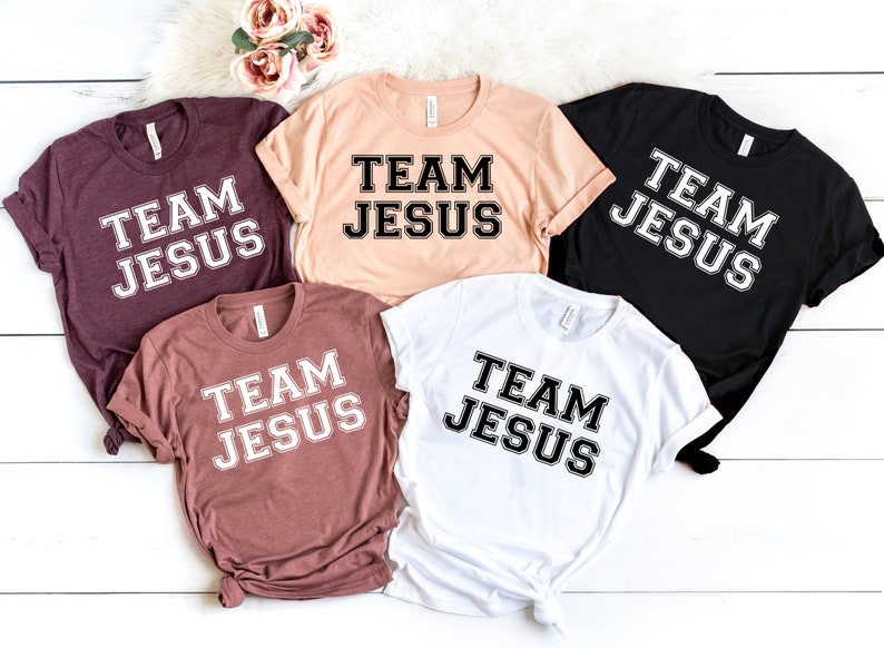 May include: Five t-shirts in different colors with the text "Team Jesus" printed in black with a white outline.