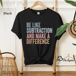 May include: Black t-shirt with the text "BE LIKE SUBTRACTION AND MAKE A DIFFERENCE" in blue, pink, and tan. The shirt is hanging on a wooden rack. The shirt is knotted at the waist.