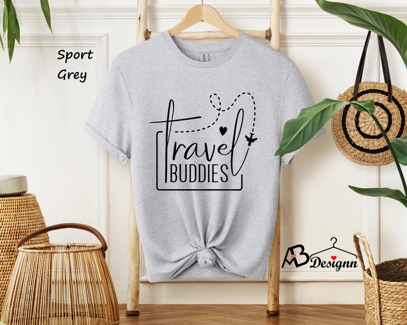 K&ouml;nnte beinhalten: Ein kurz&auml;rmeliges T-Shirt in Sportgrau mit einem schwarzen Grafikdruck, der travel BUDDIES liest, erg&auml;nzt durch eine gepunktete Flugroute, ein kleines Herz und die Silhouette eines Flugzeugs, pr&auml;sentiert auf einem Holzkleiderb&uuml;gel.
