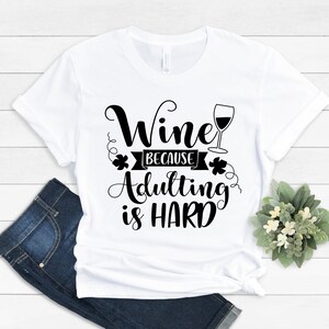 Pu&ograve; includere: Una maglietta bianca con la scritta nera "Wine because adulting is HARD". Il design include un calice di vino ed elementi decorativi. La maglietta &egrave; abbinata a jeans blu e una piccola composizione floreale.