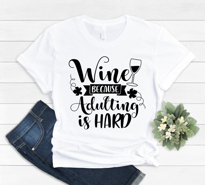 Pu&ograve; includere: Una maglietta bianca con la scritta nera "Wine because adulting is HARD". Il design include un calice di vino ed elementi decorativi. La maglietta &egrave; abbinata a jeans blu e una piccola composizione floreale.