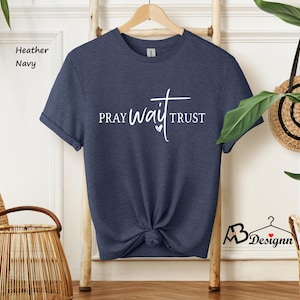 May include: Heather navy blue t-shirt with the words "PRAY WAIT TRUST" in white script. The shirt is knotted at the waist and hangs on a wooden hanger. The words are arranged with a cross above the word "wait" and a heart symbol.