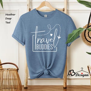 K&ouml;nnte beinhalten: Ein kurz&auml;rmeliges T-Shirt in Heather Deep Teal mit einem wei&szlig;en Grafikdruck, der travel BUDDIES liest, erg&auml;nzt durch eine gestrichelte Flugroute, ein kleines Herz und ein Flugzeugsymbol. Das T-Shirt ist in der Mitte unten zu einem Knoten gebunden.