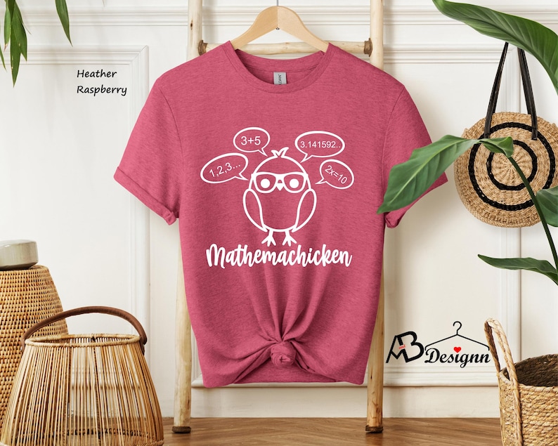 Op de afbeelding: Een heather framboos t-shirt met een witte afbeelding van een cartoonkip die een bril draagt en nadenkt over wiskundige vergelijkingen. De tekst "Mathemachicken" is onder de kip gedrukt.