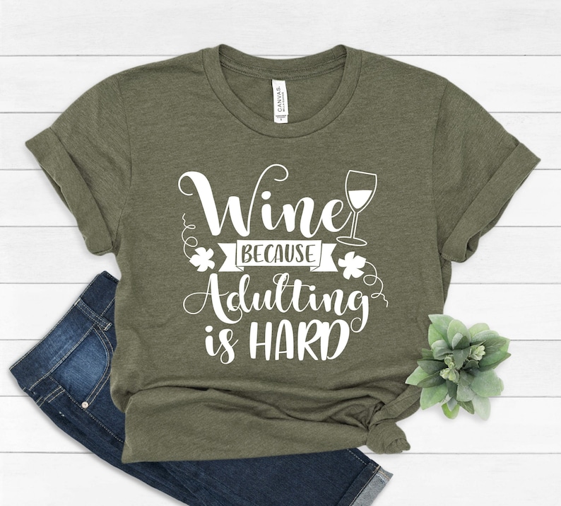 Pu&ograve; includere: T-shirt verde oliva con la scritta bianca "Wine because Adulting is HARD". Il design include un calice e decorazioni. La maglietta &egrave; piegata con un paio di jeans.