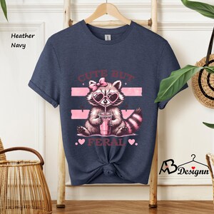 Op de afbeelding: Een heather navy blauw t-shirt met een afbeelding van een wasbeer die een zonnebril draagt en een roze drankje drinkt. De tekst "Cute But Feral" staat op het shirt gedrukt.
