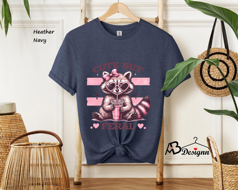 Op de afbeelding: Een heather navy blauw t-shirt met een afbeelding van een wasbeer die een zonnebril draagt en een roze drankje drinkt. De tekst "Cute But Feral" staat op het shirt gedrukt.