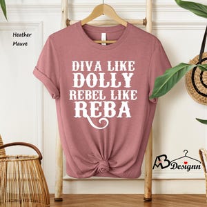 Puede incluir: Una camiseta de manga corta en color malva jaspeado con texto blanco que dice DIVA LIKE DOLLY REBEL LIKE REBA. La camiseta tiene cuello redondo y se muestra en una percha de madera. La tela tiene un aspecto jaspeado suave con un dobladillo anudado.