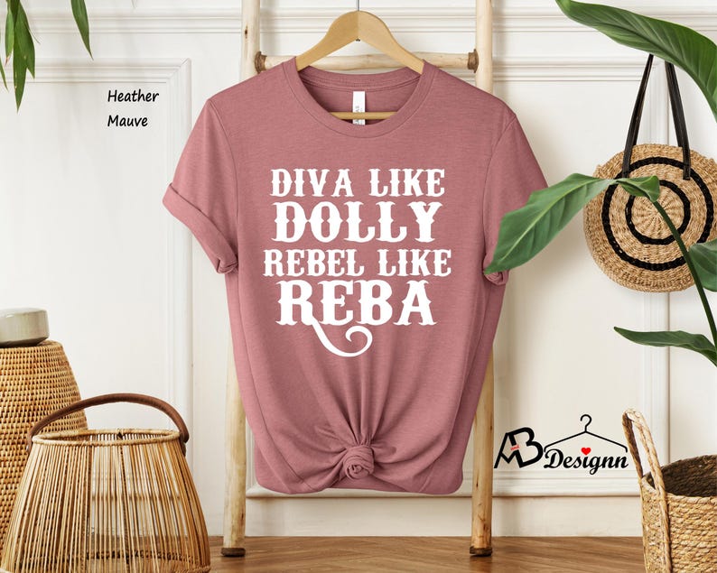Puede incluir: Una camiseta de manga corta en color malva jaspeado con texto blanco que dice DIVA LIKE DOLLY REBEL LIKE REBA. La camiseta tiene cuello redondo y se muestra en una percha de madera. La tela tiene un aspecto jaspeado suave con un dobladillo anudado.