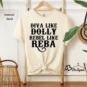 Puede incluir: Una camiseta de manga corta en color arena natural con texto negro que dice DIVA LIKE DOLLY REBEL LIKE REBA. La prenda tiene cuello redondo y est&aacute; colgada en una percha de madera. La tela es de un tono beige claro s&oacute;lido con letras.