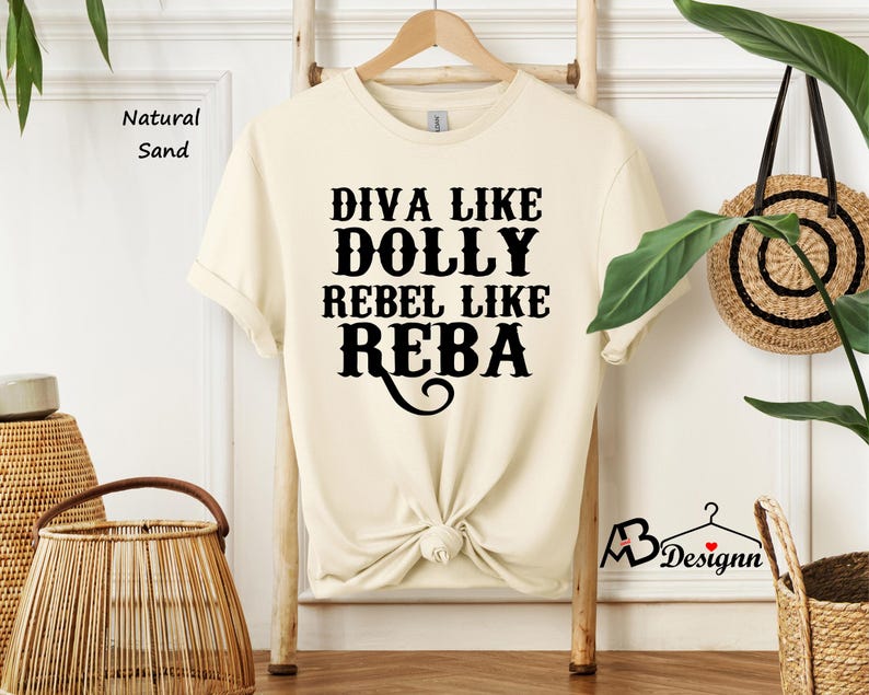Puede incluir: Una camiseta de manga corta en color arena natural con texto negro que dice DIVA LIKE DOLLY REBEL LIKE REBA. La prenda tiene cuello redondo y est&aacute; colgada en una percha de madera. La tela es de un tono beige claro s&oacute;lido con letras.