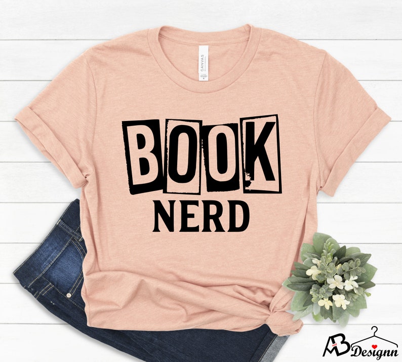 Może przedstawiać: Jasnor&oacute;żowy t-shirt z napisem "BOOK NERD" czarnymi literami. Litery są w stylu vintage i zużytym.