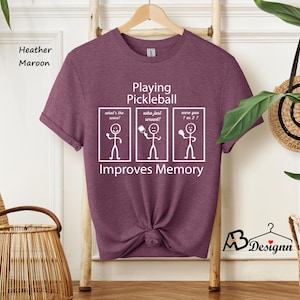 May include: A heather maroon t-shirt with a white graphic that says "Playing Pickleball Improves Memory". The graphic features three stick figures with paddles, each with a caption: "What's the score?", "Who just served?", and "Were you 7 or 2?"