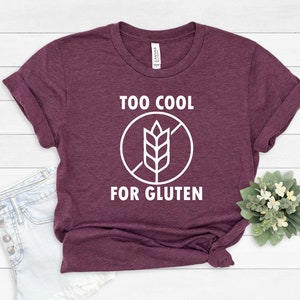 May include: A maroon t-shirt with white text that reads "Too Cool For Gluten" and a white graphic of a wheat stalk with a diagonal line through it.
