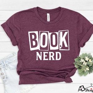 Może przedstawiać: Bordowy t-shirt z nadrukiem "BOOK NERD" w białych literach. Litery są w blokowym kroju i wyglądają na wycięte z papieru.
