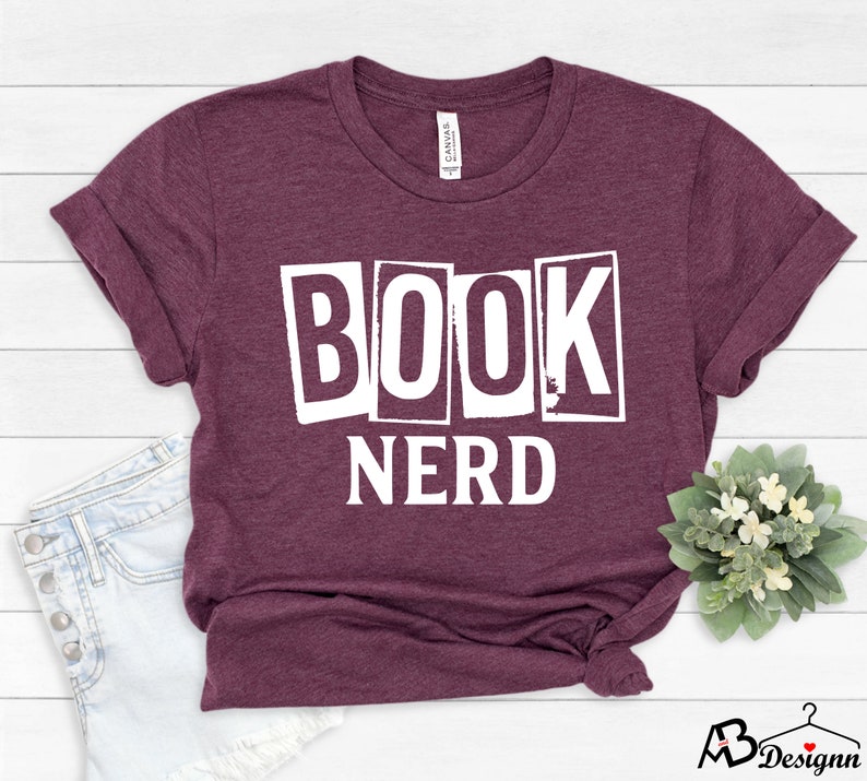 Może przedstawiać: Bordowy t-shirt z nadrukiem "BOOK NERD" w białych literach. Litery są w blokowym kroju i wyglądają na wycięte z papieru.