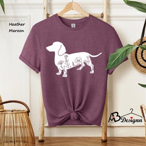 May include: A heather maroon t-shirt featuring a white outline of a dachshund with floral details. The shirt is on a hanger and has a knotted bottom.