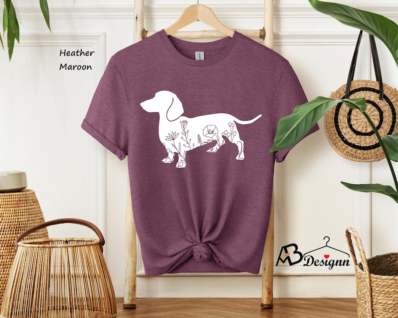 May include: A heather maroon t-shirt featuring a white outline of a dachshund with floral details. The shirt is on a hanger and has a knotted bottom.