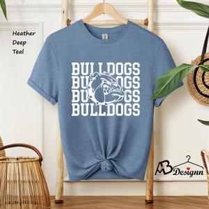 Puede incluir: Camiseta de color verde azulado jaspeado con la palabra "BULLDOGS" apilada verticalmente en letras blancas. Un gr&aacute;fico de bulldog blanco est&aacute; centrado en la camiseta. La camiseta est&aacute; colgada de una percha de madera.