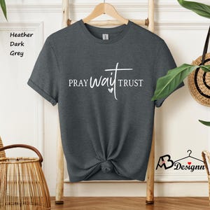 May include: Heather dark grey t-shirt with the words "PRAY WAIT TRUST" in white script. The shirt is knotted at the waist and hangs on a wooden hanger. The shirt is displayed in a well-lit room.