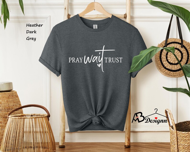 May include: Heather dark grey t-shirt with the words "PRAY WAIT TRUST" in white script. The shirt is knotted at the waist and hangs on a wooden hanger. The shirt is displayed in a well-lit room.