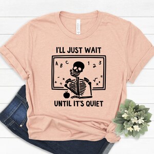 Może przedstawiać: R&oacute;żowy t-shirt z czarną ilustracją szkieletu. Szkielet siedzi przy tablicy z napisem "Po prostu poczekam, aż będzie cicho".