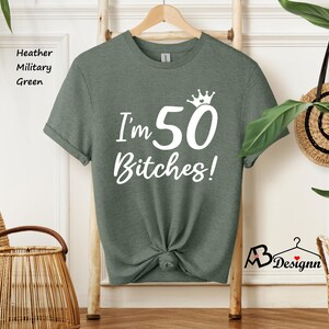 May include: A heather military green short-sleeve t-shirt featuring white text that reads I'm 50 Bitches! with a small crown graphic above the number 50. The shirt has a crew neckline and is displayed on a wooden hanger against a light wall.