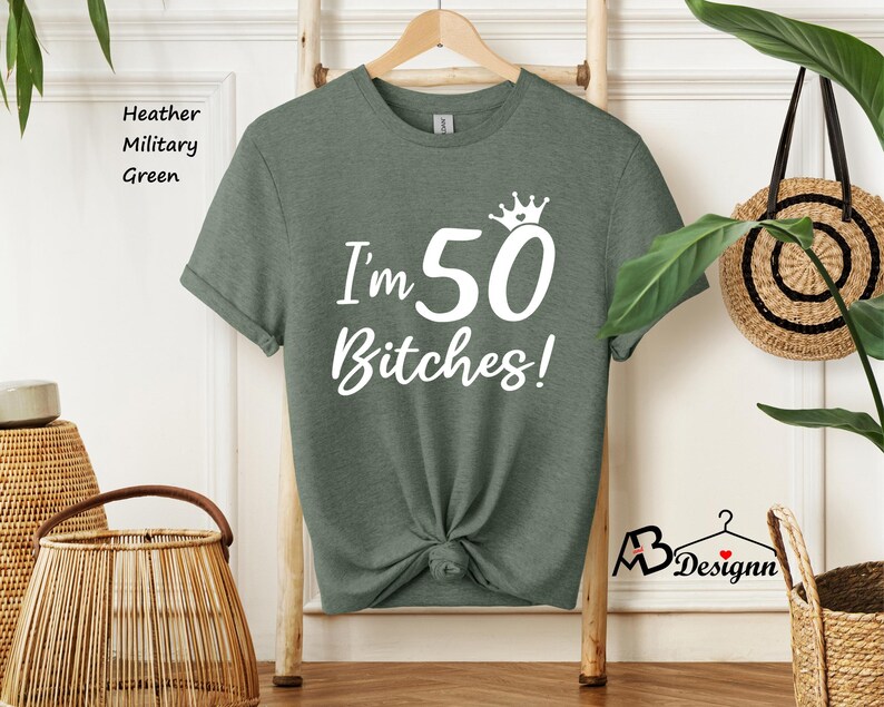 May include: A heather military green short-sleeve t-shirt featuring white text that reads I'm 50 Bitches! with a small crown graphic above the number 50. The shirt has a crew neckline and is displayed on a wooden hanger against a light wall.