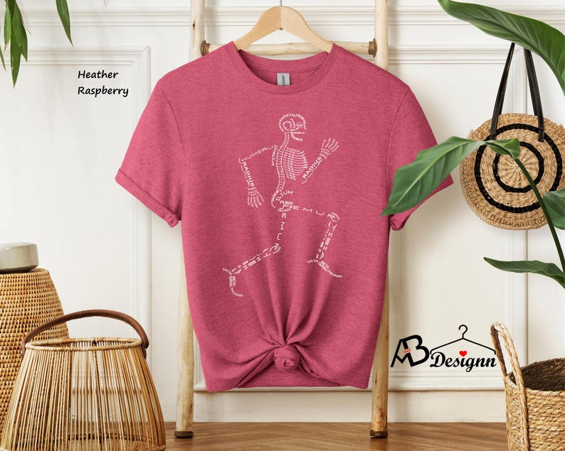 May include: Heather raspberry t-shirt featuring a white skeleton running with the names of the bones labeled. The shirt is on a wooden hanger.