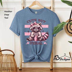 Op de afbeelding: Een heide diep teal t-shirt met een afbeelding van een wasbeer die een hartvormige zonnebril draagt en drinkt uit een roze beker. De tekst "Cute But Feral" is op het shirt gedrukt.