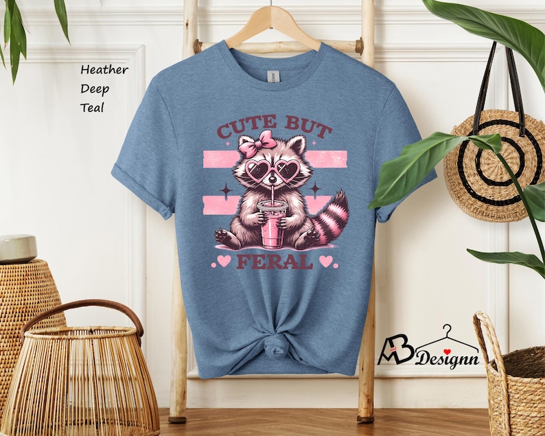 Op de afbeelding: Een heide diep teal t-shirt met een afbeelding van een wasbeer die een hartvormige zonnebril draagt en drinkt uit een roze beker. De tekst "Cute But Feral" is op het shirt gedrukt.