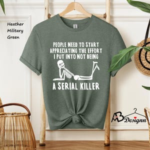 May include: A heather military green t-shirt with a white skeleton graphic and the text "People need to start appreciating the effort I put into not being a serial killer".
