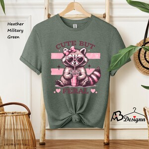 Op de afbeelding: Een heide-militaire groene t-shirt met een afbeelding van een wasbeer die een zonnebril draagt en een roze drankje vasthoudt. De tekst "Cute But Feral" is onder de wasbeer gedrukt.