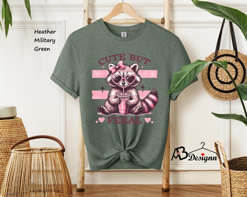 Op de afbeelding: Een heide-militaire groene t-shirt met een afbeelding van een wasbeer die een zonnebril draagt en een roze drankje vasthoudt. De tekst "Cute But Feral" is onder de wasbeer gedrukt.