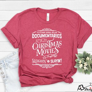 May include: A maroon t-shirt with white text that reads "It's Either Serial Killer Documentaries or Christmas Movies, We Either Sleighin' or Slayin'!" The shirt is folded, with a small plant and a pair of jeans in the background.