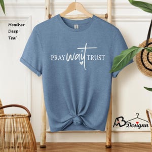 May include: A heather deep teal t-shirt with the words "PRAY WAIT TRUST" in white script and a cross design. The shirt is displayed on a wooden hanger against a white wall.